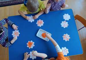 Dzieci siedzące przy stoliku. Widziane z góry. Stemplują kwiatki wycięte z papieru przy użyciu patyczków higienicznych.